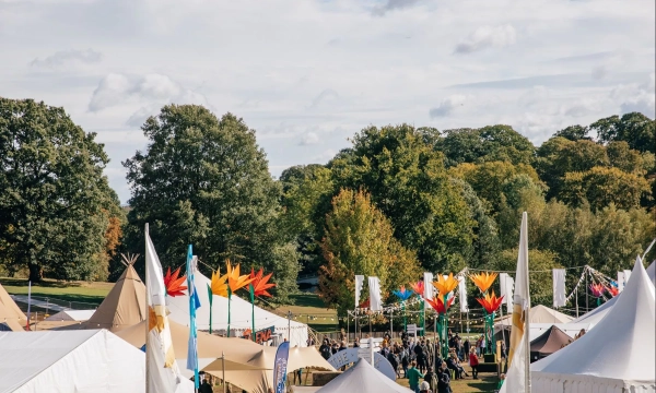 A vibrant outdoor festival scene featuring colorful decorations, tents, and people mingling among trees under a partly cloudy sky.