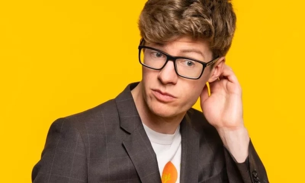 A young man in a gray blazer and glasses looks over his shoulder, standing against a bright yellow background.