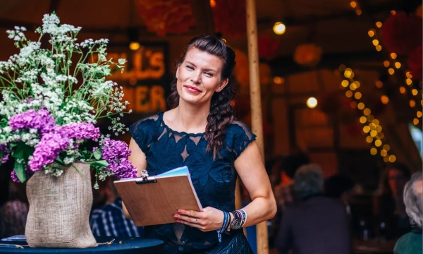 A smiling woman with long braided hair holds a clipboard near a floral arrangement in a festive indoor setting.