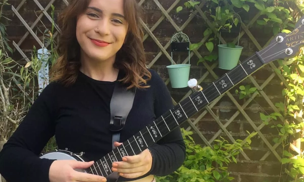 A smiling young woman holds a banjo, standing in a green garden with climbing plants and hanging pots in the background.