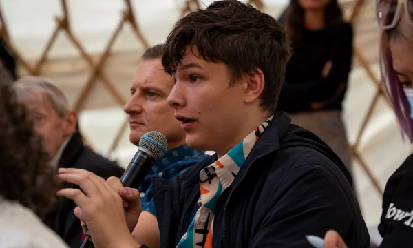 A young man with curly hair seated in a tent, speaking into a microphone while engaging with an audience. Other attendees are visible in the background.