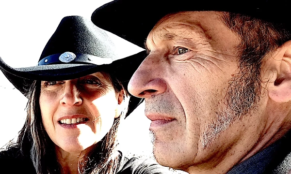 A man and woman wearing black cowboy hats pose for a selfie against a bright background, both showcasing strong facial features and expressions.