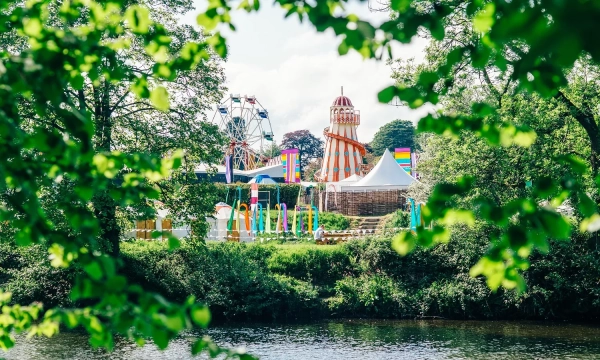 A vibrant amusement park scene featuring a carousel, a Ferris wheel, and colorful tents, framed by green foliage beside a tranquil river.