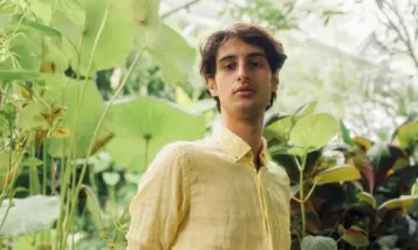 A young man with long hair stands amidst lush greenery in a greenhouse, wearing a yellow shirt and posing confidently.