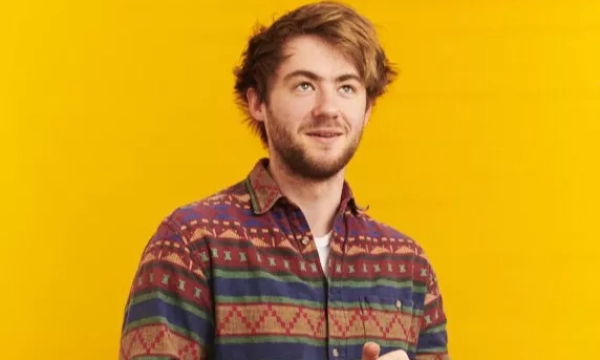 A young man with tousled hair, wearing a colorful patterned shirt, poses against a vibrant yellow background.