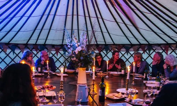 A cozy gathering inside a yurt, with a large round table, candlelight, and guests engaged in conversation amidst floral decorations.