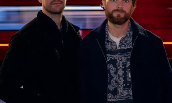 Two men stand in front of a blurred red and blue background, with a city street visible at night. One man wears a cap and a black jacket, while the other sports a patterned shirt.
