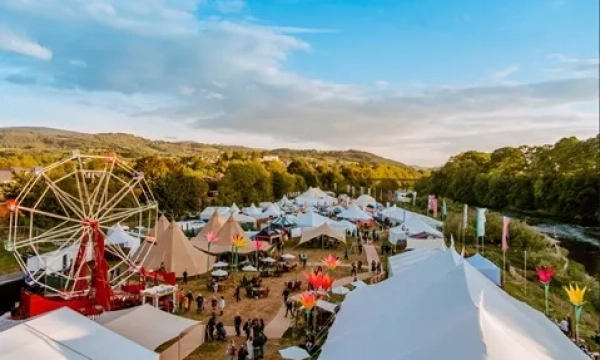 A vibrant outdoor festival setting featuring tents, a Ferris wheel, and attendees enjoying the festivities along a riverbank, surrounded by lush greenery and scenic hills.