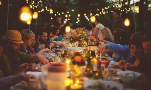 A long outdoor dining table set for a communal meal, surrounded by softly glowing string lights and people enjoying their evening with food and conversation.