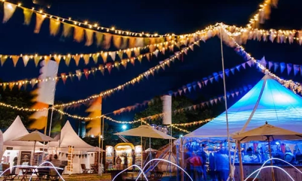 A vibrant outdoor festival scene at night, adorned with colorful bunting and string lights, featuring tents and people socializing.