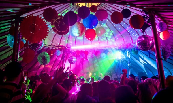 A vibrant indoor party scene with colorful decorations, disco balls, and dynamic lighting, filled with people dancing under a fabric canopy.