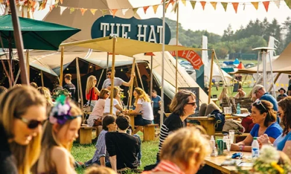 A lively outdoor festival scene with colorful tents and people enjoying food and drinks, seated at wooden tables, surrounded by vibrant bunting and greenery.