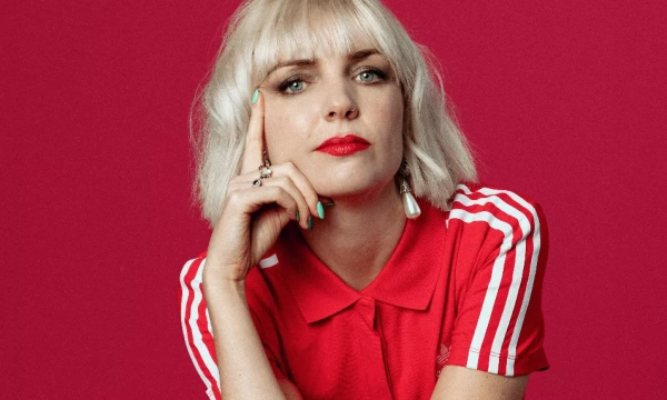 A woman with short blonde hair, wearing a red polo shirt with white stripes, poses against a vibrant pink background, looking thoughtfully at the camera.