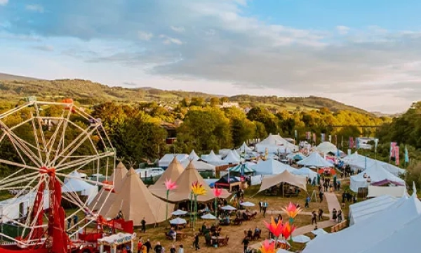 A vibrant outdoor festival scene featuring colorful tents, a large Ferris wheel, and decorated areas amidst lush green hills and trees under a blue sky.