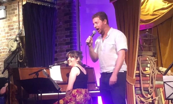 A man and a woman perform on stage, with the woman at a piano and the man singing into a microphone, set against a vibrant backdrop.