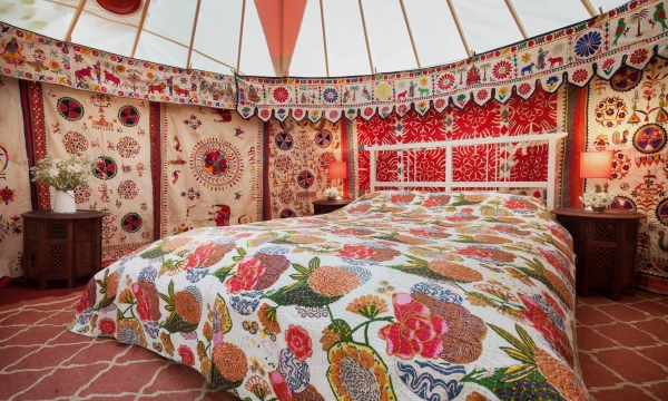 A beautifully decorated yurt interior featuring a vibrant, floral-patterned quilt on a bed, adorned with colorful wall hangings and ambient lighting.