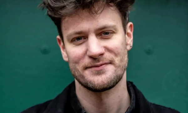 A man with tousled hair smiles at the camera, wearing a black jacket against a green background.