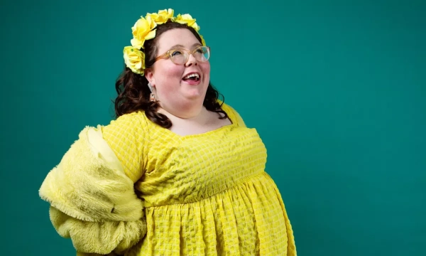 A cheerful individual with curly hair and glasses, wearing a yellow dress and a flower crown, smiles brightly against a teal background.