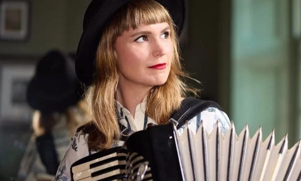 A woman with long hair, wearing a hat and a floral shirt, plays an accordion, looking thoughtfully to the side.