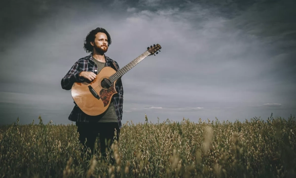 A man stands in a field holding an acoustic guitar, with a cloudy sky overhead and tall grass surrounding him.