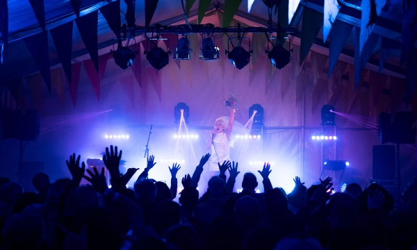 A vibrant concert scene featuring a performer with arms raised, engaging an audience with hands lifted, all under colorful bunting and stage lights in a festive environment.