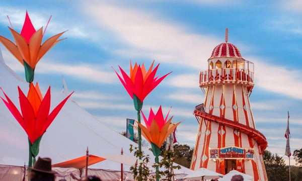 A vibrant carnival scene featuring a colorful slide tower surrounded by decorative large flowers and tents against a cloudy sky.