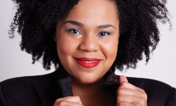 A smiling woman with curly black hair, wearing a striking black and red jacket, exudes confidence and warmth against a neutral background.