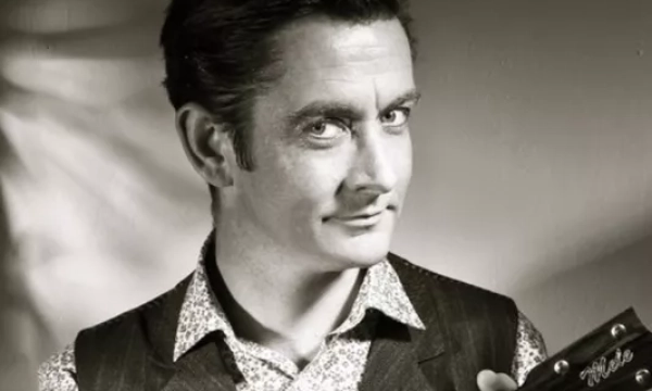 A charismatic man with a mustache and slicked-back hair, wearing a patterned shirt and vest, holds a ukulele, evoking a vintage musical vibe.