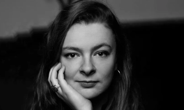 A close-up portrait of a woman with long, wavy hair, resting her chin on her hand, and looking directly at the camera with a thoughtful expression.