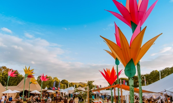 Colorful oversized flower decorations in a festival setting, with tents and crowds of people enjoying the event under a bright blue sky.