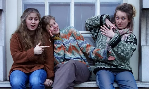 Three young women sitting on a ledge, wearing colorful sweaters, making playful expressions and poses, showcasing a sense of friendship and fun.