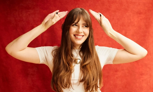Woman with long hair and a bright smile, playfully posing with her hands raised over her head against a red background.