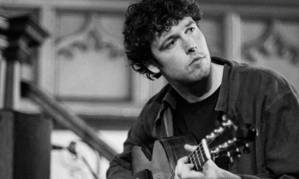 A musician with curly hair plays an acoustic guitar, focused and immersed in the music, against a softly lit backdrop.