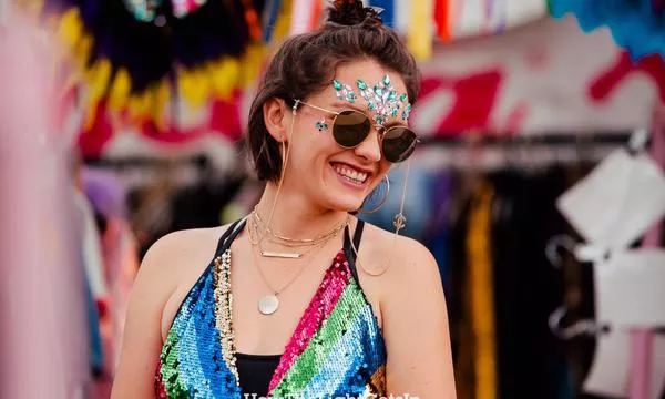 A smiling young woman wearing a colorful sequin outfit, sunglasses, and decorative jewels on her forehead, standing in a vibrant festival environment.